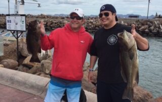 Two men each holding a fresh fish standing on a road in front of ocean